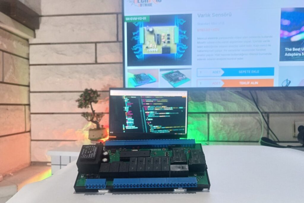 ön planda üzerinde elektronik parçalar olan TNS online parosyonel varlık modülü devresi, arka planda taş duvarda asılı televizyon ve altında ekranında yazılım kodları olan laptop görseli