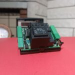 Arka planda taş duvarlı şömine ve saat, ön planda kırmızı zemin üstünde üzerinde elektronik kompenentler olan TNS online varlık modülü lite devresi olan görsel