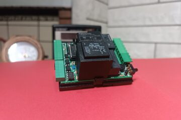 Arka planda taş duvarlı şömine ve saat, ön planda kırmızı zemin üstünde üzerinde elektronik kompenentler olan TNS online varlık modülü lite devresi olan görsel