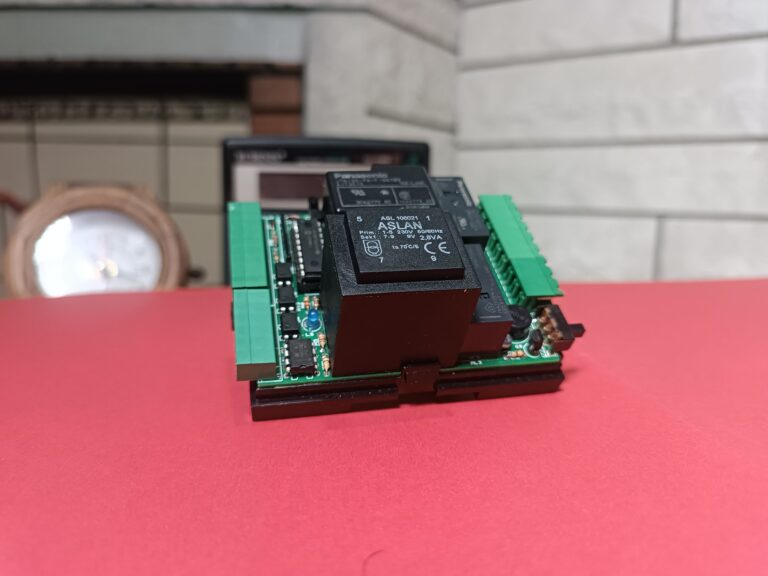 Arka planda taş duvarlı şömine ve saat, ön planda kırmızı zemin üstünde üzerinde elektronik kompenentler olan TNS online varlık modülü lite devresi olan görsel
