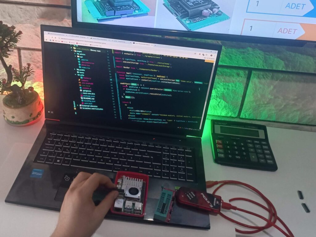 tezgah üzerinde laptop, ekranında kodlama sayfası olan ve arkada tv ekranında varlık modülleri olan görsel